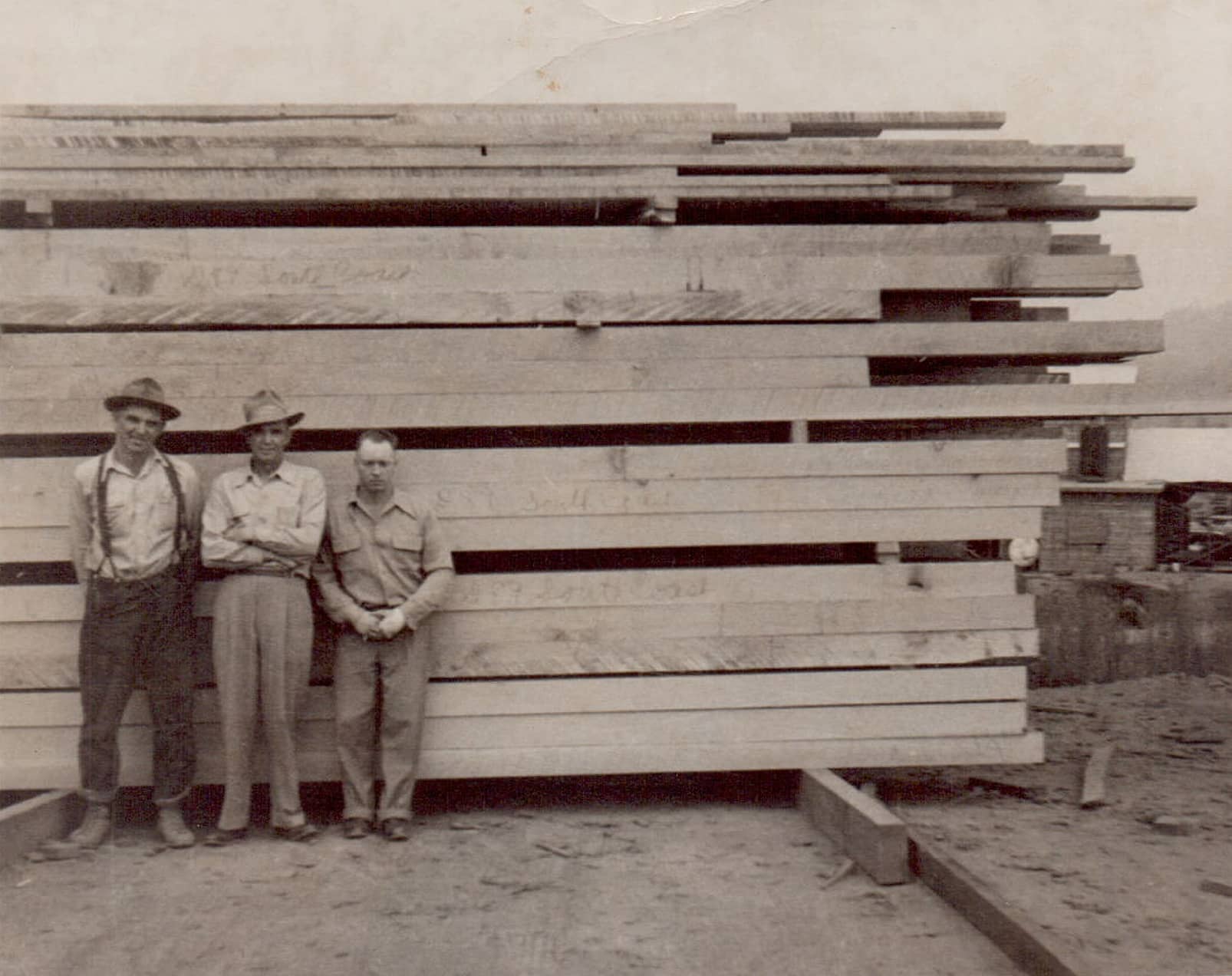 Bill, Archie, and Charlie stand in front of a stack of lumber